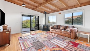 A spacious living area with a wooden ceiling, a large sofa, a fireplace, and a view through a sliding glass door. - Ocean Views on Aldebaran - sleeps 11 (Ocean Grove)