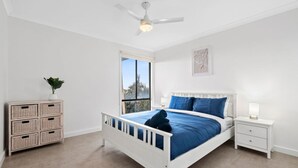 A bedroom with a white bed, a nightstand, a dresser, a ceiling fan, and a window. - Ocean Views on Aldebaran - sleeps 11 (Ocean Grove)