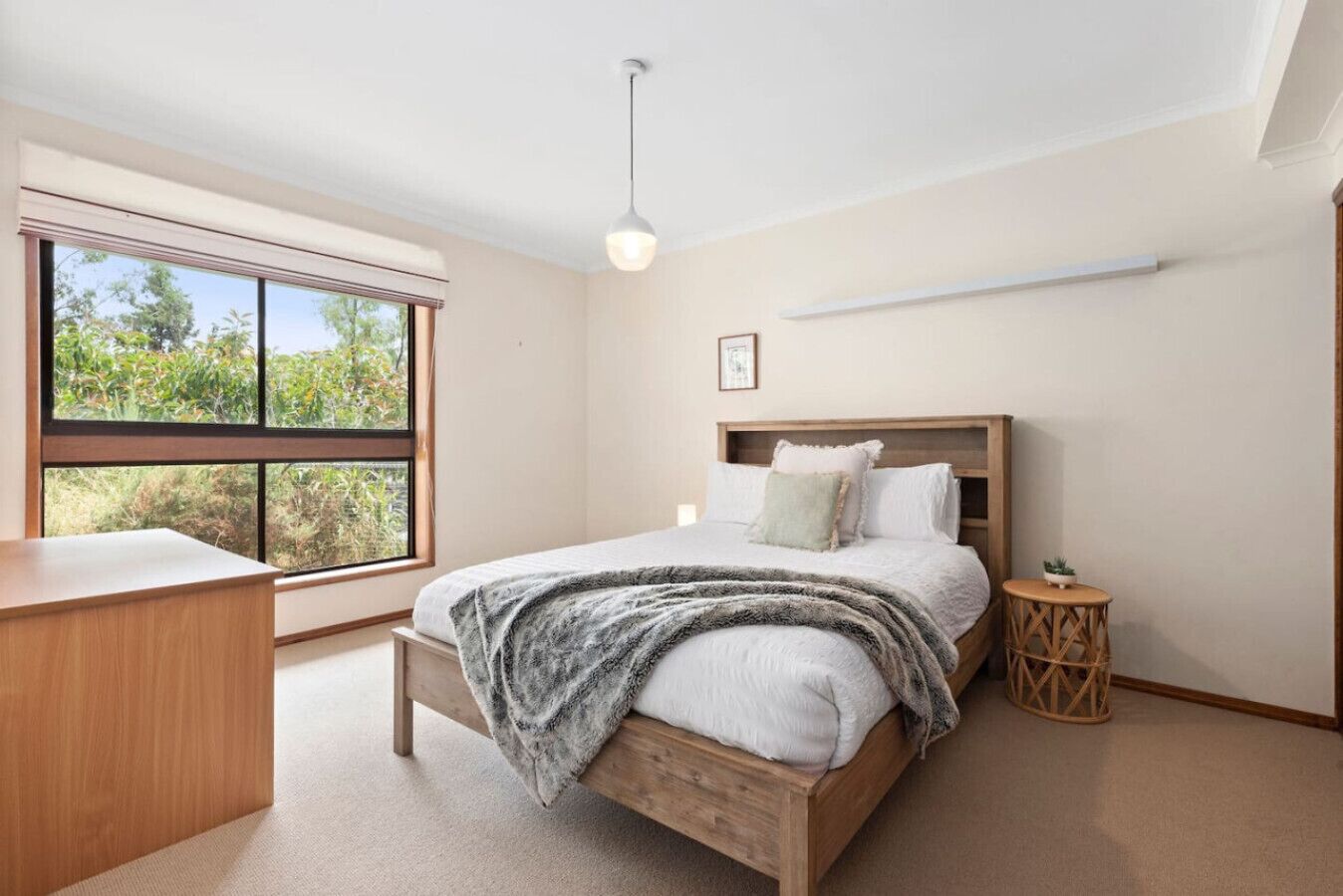 Une chambre avec un lit en bois, un bureau, une table de chevet, une lampe suspendue et une fenêtre donnant sur un paysage verdoyant.