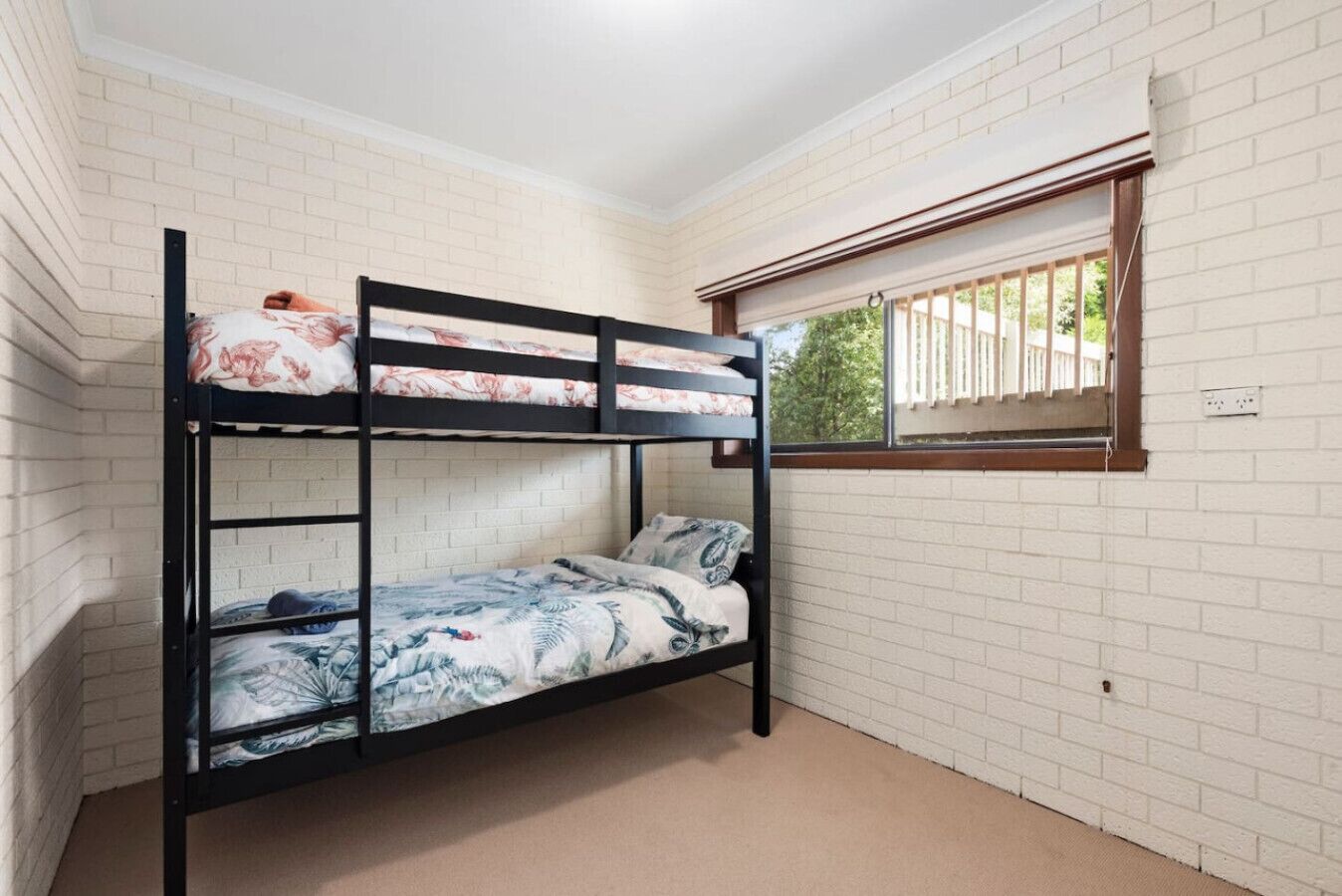 Een slaapkamer met stapelbedden, witte bakstenen muren, een raam met zonwering en een zichtbaar stopcontact.