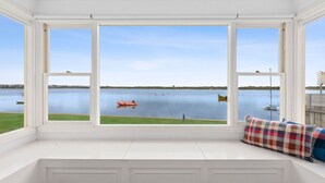 Ein Zimmer mit einem großen Erkerfenster mit Blick auf ruhiges Wasser, ein kleines Boot und eine Rasenfläche.