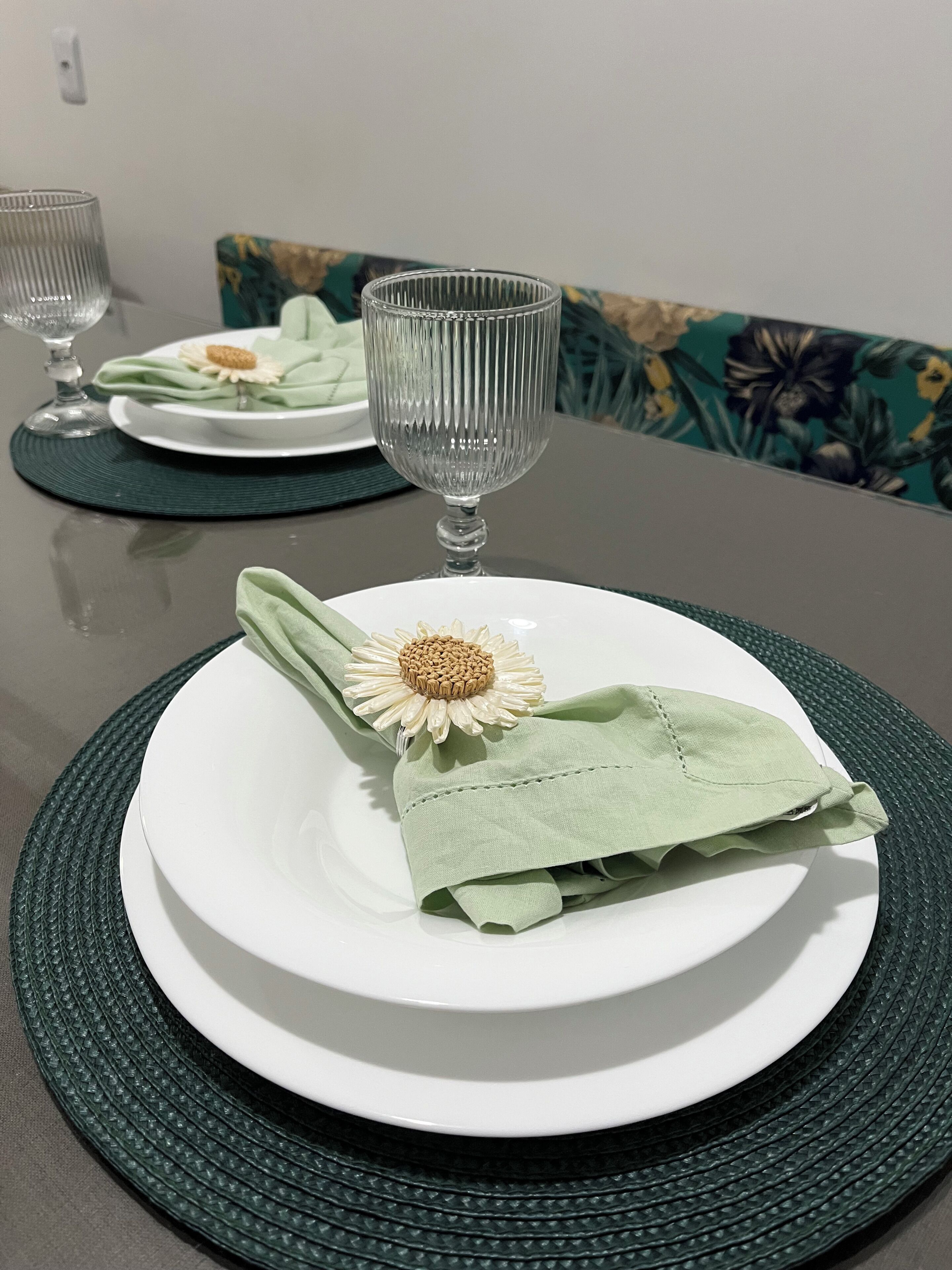 A dining table with a white plate, a green napkin, a glass, and a floral cushion in the background.