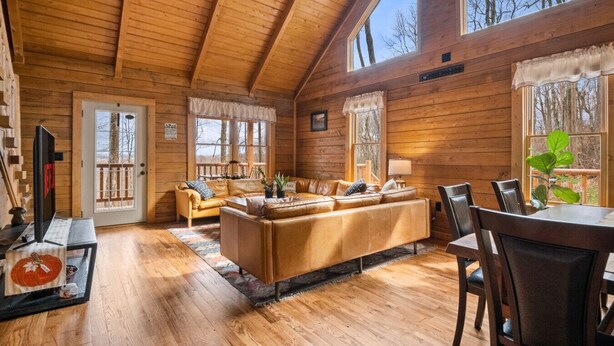 A log cabin interior with a sofa set, dining table, and chairs, large windows, and a wooden ceiling.