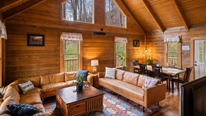 A log cabin interior with a wooden ceiling, large windows, a sofa, a coffee table, and a dining area.
