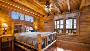 A log cabin bedroom with a bed, nightstands, a ceiling fan, and large windows.
