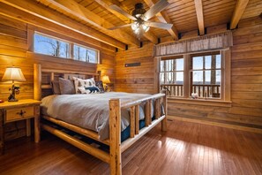 Un dormitorio tipo cabaña de troncos con una cama, mesitas de noche, ventilador de techo y ventanas amplias.