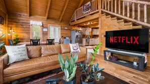 A cozy cabin interior with a wooden staircase, a living room with a leather sofa, a kitchen area, and a TV displaying 'NETFLIX'.