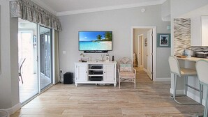 Un salon comprenant un téléviseur, un meuble de rangement blanc, un fauteuil et un coin repas avec des tabourets de bar.