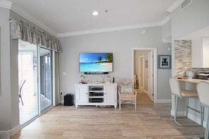 Sala de estar con televisor, mueble blanco para entretenimiento, una silla y comedor con taburetes de bar.
