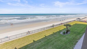 A beach view with a grassy area, a palm tree, and a fence. - Oceanview Studio w/ Private Balcony, Pools & Beach Access (Daytona Beach Shores)