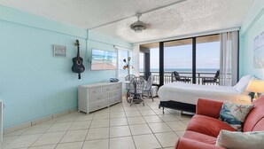 A room with a bed, a red couch, a guitar, a radiator, a table with chairs, and a view of the ocean. - Oceanview Studio w/ Private Balcony, Pools & Beach Access (Daytona Beach Shores)