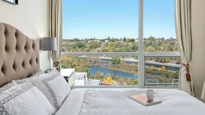 Een hotelkamer met een groot raam, een bed, een nachtkastje en uitzicht op een meer en gebouwen.