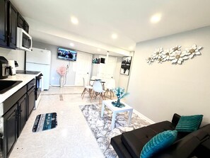 A modern kitchen with a microwave, stove, and sink. There is a dining area with a white table and chairs. A black sofa with teal cushions is in the living area.