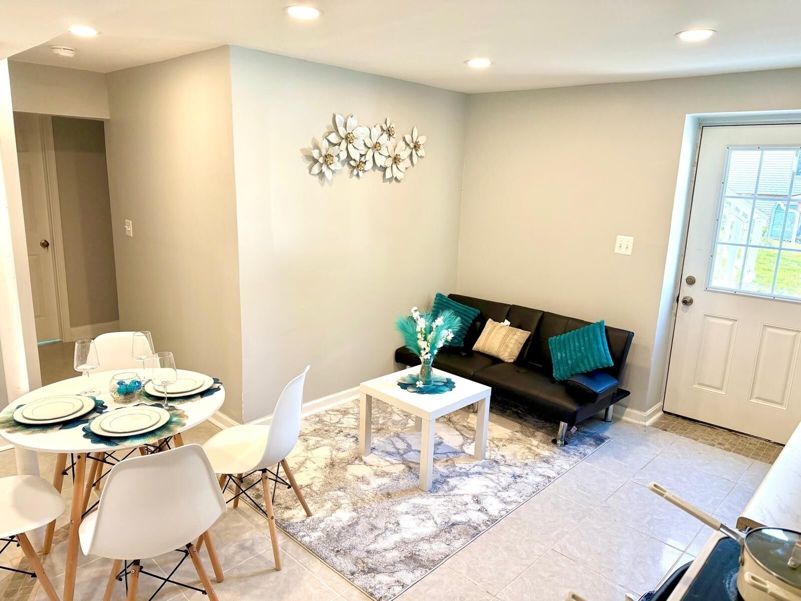 A dining area with a round table, white chairs, and a vase with flowers. There is a black sofa with teal cushions and a decorative wall piece.