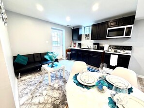 A modern living room with a sofa, coffee table, and dining table set for six. A kitchen with dark cabinets and a microwave is visible in the background.