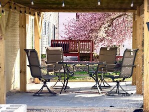 An outdoor patio with a table and four chairs, a wooden pergola, and a view of a cherry blossom tree.