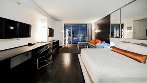 Una habitación de hotel moderna con una cama grande, un escritorio con televisor y vistas a la ciudad desde las ventanas.