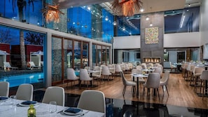 A modern dining area with multiple tables and chairs, large windows, and a pool view.