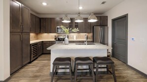 A modern kitchen with dark wood cabinets, a central island with stools, stainless steel appliances, and pendant lighting. - Landing | Stunning 2BD, Gym, Game Room (Fort Worth)