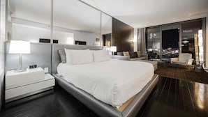 Ett modernt hotellrum med en stor säng, ett nattduksbord, en lampa och utsikt över staden på natten.