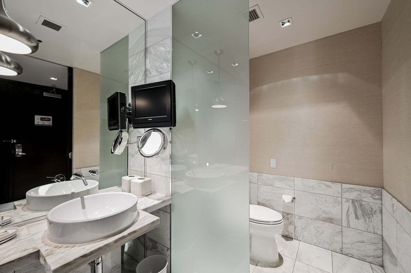 Et moderne badeværelse med to håndvaske, toilet og et stort spejl med indbygget tv.
