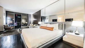 Ein modernes Hotelzimmer mit einem großen Bett, einer Sitzecke mit Sofa und Sesseln, einem Nachttisch mit Lampe und Blick auf die Stadt durch das Fenster.