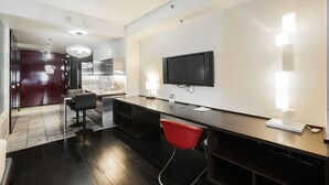 Ein modernes Hotelzimmer mit dunklem Holzboden, einem schwarzen Schreibtisch, einem roten Sessel, einem großen Spiegel, einem Flachbildfernseher und einer wandmontierten Leuchte.