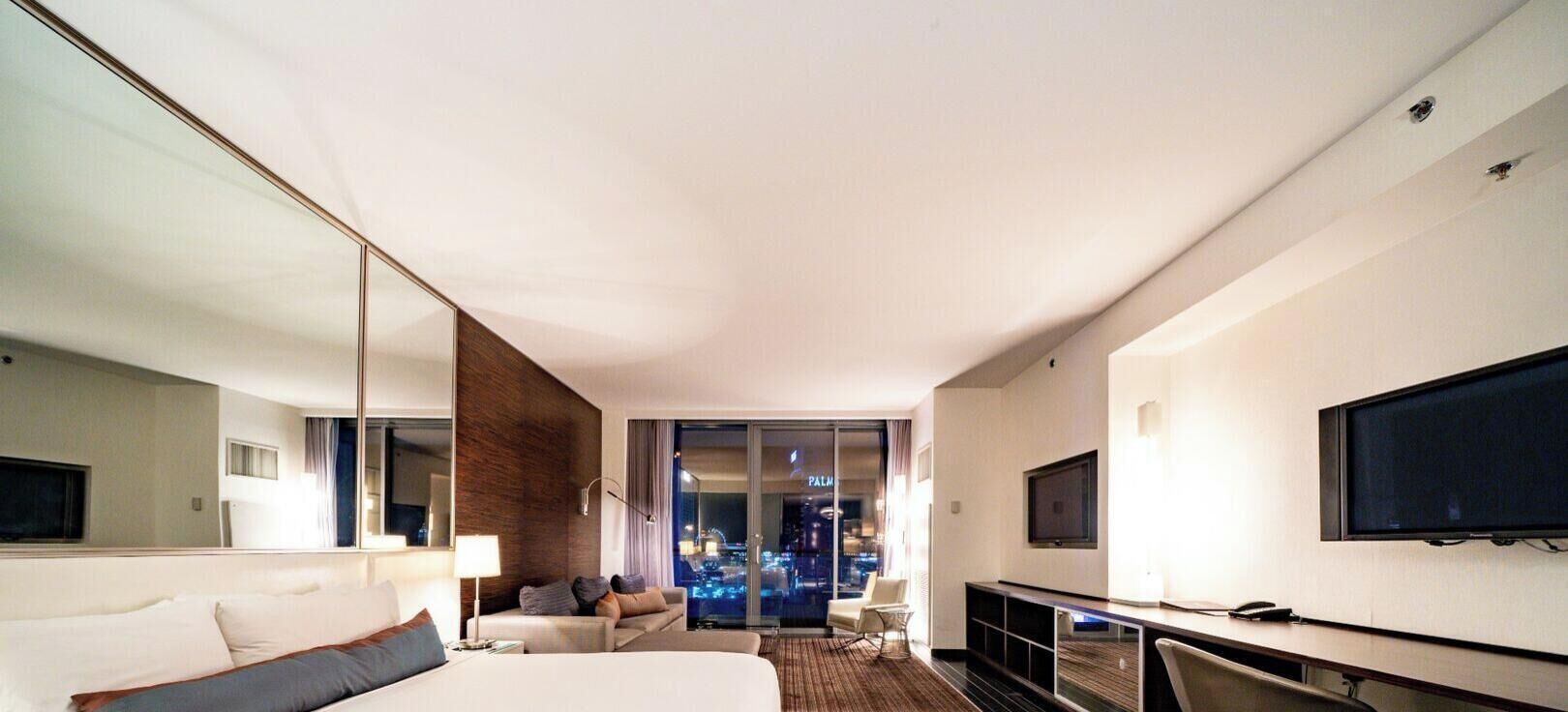 Ein modernes Hotelzimmer mit einem großen Bett, einem Schreibtisch mit Fernseher, einer Sitzecke und Blick auf eine Stadtlandschaft.