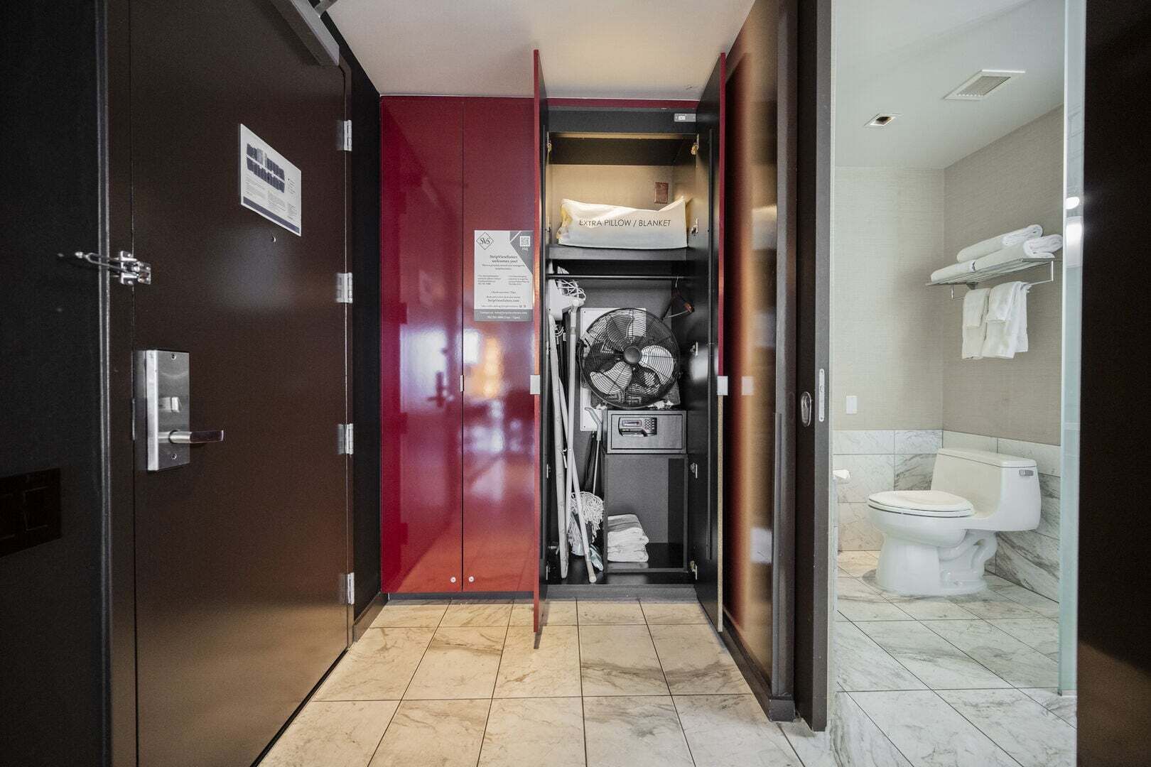 Een hotelkamer met een rode deur, een wit toilet, een ventilator en een handdoekhouder.