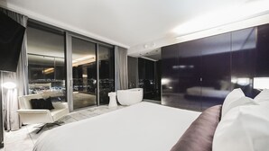 Et moderne hotelværelse med en stor seng, siddeplads og udsigt over byen gennem gulv til loft-vinduer.