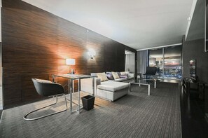 Ein modernes Hotelzimmer mit dunkler Holzvertäfelung, einer Couch, einem kleinen Tisch, einem Stuhl und Blick auf die beleuchtete Stadt bei Nacht.