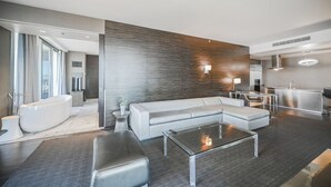 Ein modernes Wohnzimmer mit einem Ecksofa, einem Glastisch, einem großen Fenster und einem separaten Badezimmerbereich.