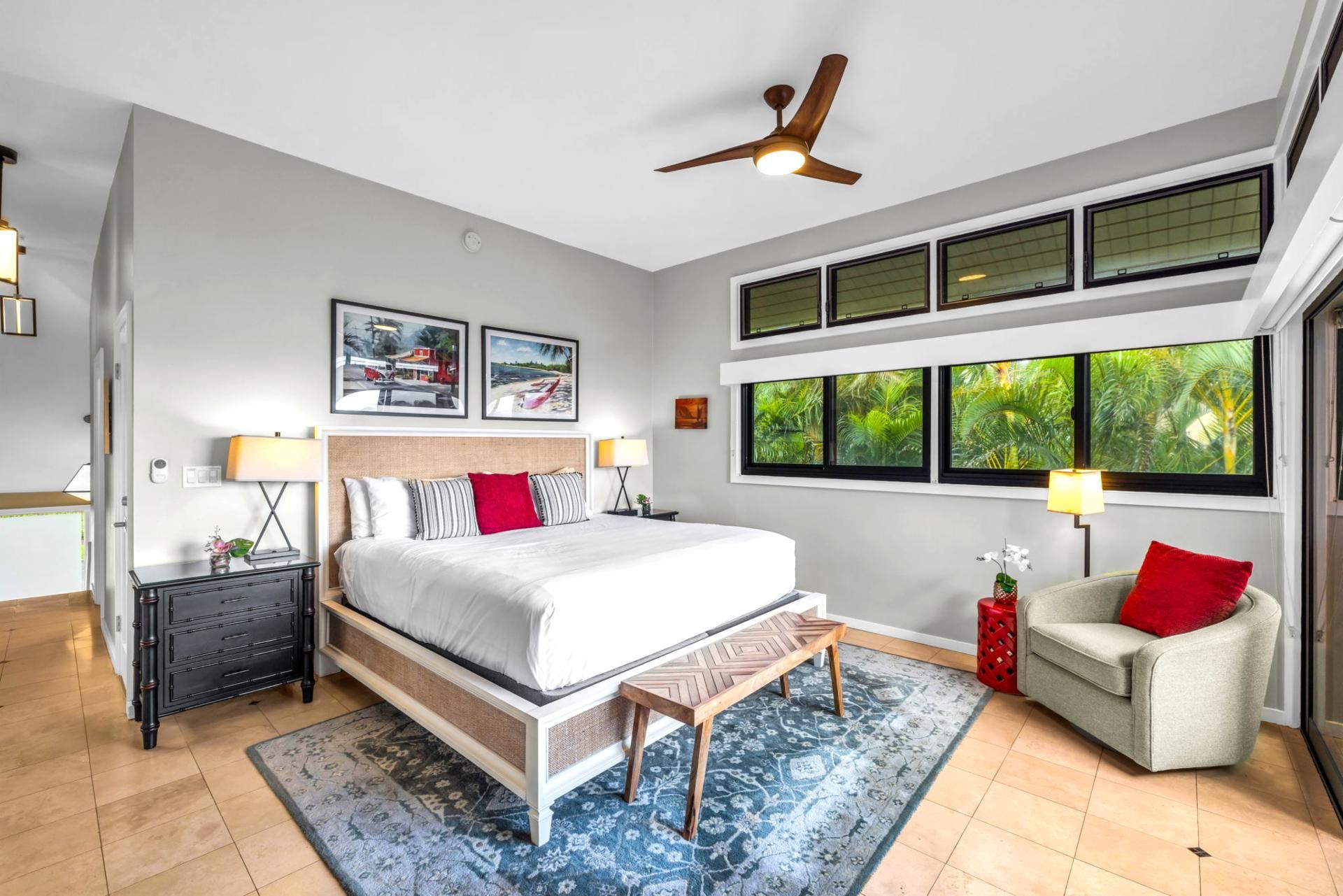 A bedroom with a bed, a nightstand, a chair, a ceiling fan, and large windows with a view of greenery.