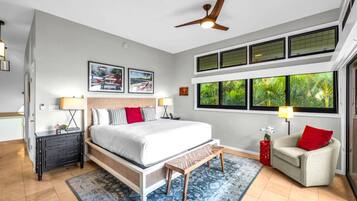 A bedroom with a bed, a nightstand, a chair, a ceiling fan, and large windows with a view of greenery.