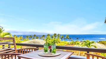 A wooden deck with a table and chairs, overlooking a beach with palm trees and the ocean.