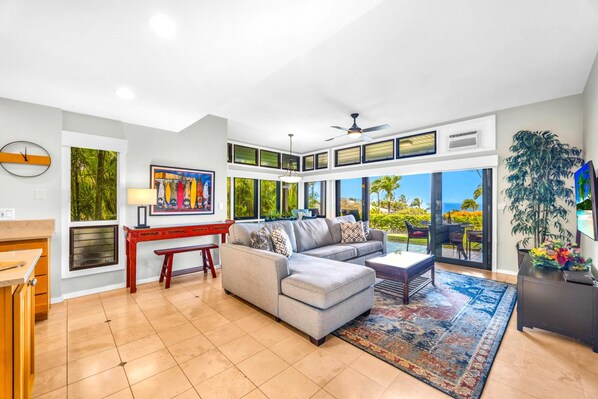 A spacious living room with a large gray sectional sofa, a wooden coffee table, a red bench, and a large window offering a view of the outdoors.