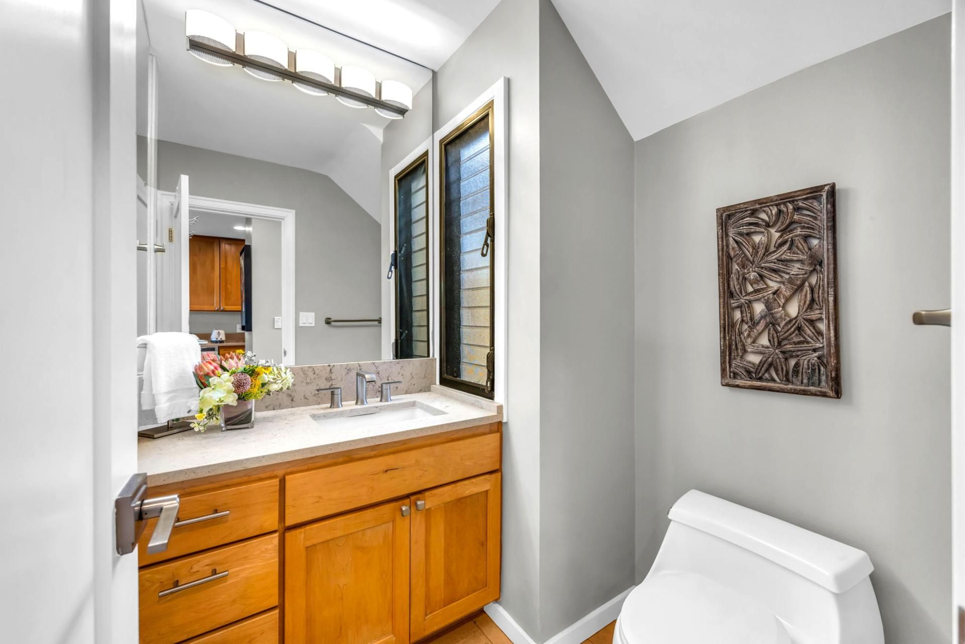 A bathroom with a white toilet, wooden cabinets, a large mirror, and a wall-mounted artwork.