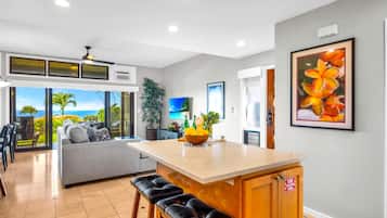 A modern kitchen with a central island, a dining area with a view, and a living space with a sofa and a TV.