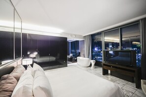 A modern hotel room with a large bed, a seating area, a bathtub, a television, and a city view. - SVS: 1BD Strip View | Jetted Tub | Full Kitchen | Sleeps 4 | Palms Place (Las Vegas)