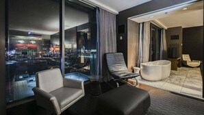 Una habitación de hotel moderna con vistas a la ciudad, un ventanal grande, un sillón de cuero negro, una bañera blanca y un área de descanso con una butaca y reposapiés.