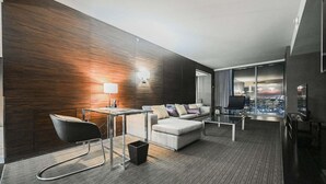 Ein modernes Hotelzimmer mit dunkler Holzvertäfelung, einer Couch, einem kleinen Tisch, einem Stuhl und Blick auf die beleuchtete Stadt bei Nacht.