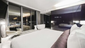 Ein modernes Hotelzimmer mit einem großen Bett, einer Sitzecke mit Stuhl und Sofa, einem großen Fenster mit Blick auf die Stadt und einer Glasschiebetür zu einem Außenbereich.