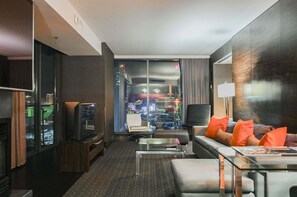 Ein modernes Hotelzimmer mit Blick auf die Stadt, einem Flachbildfernseher, einer Couch mit orangefarbenen Kissen und einem Glastisch.