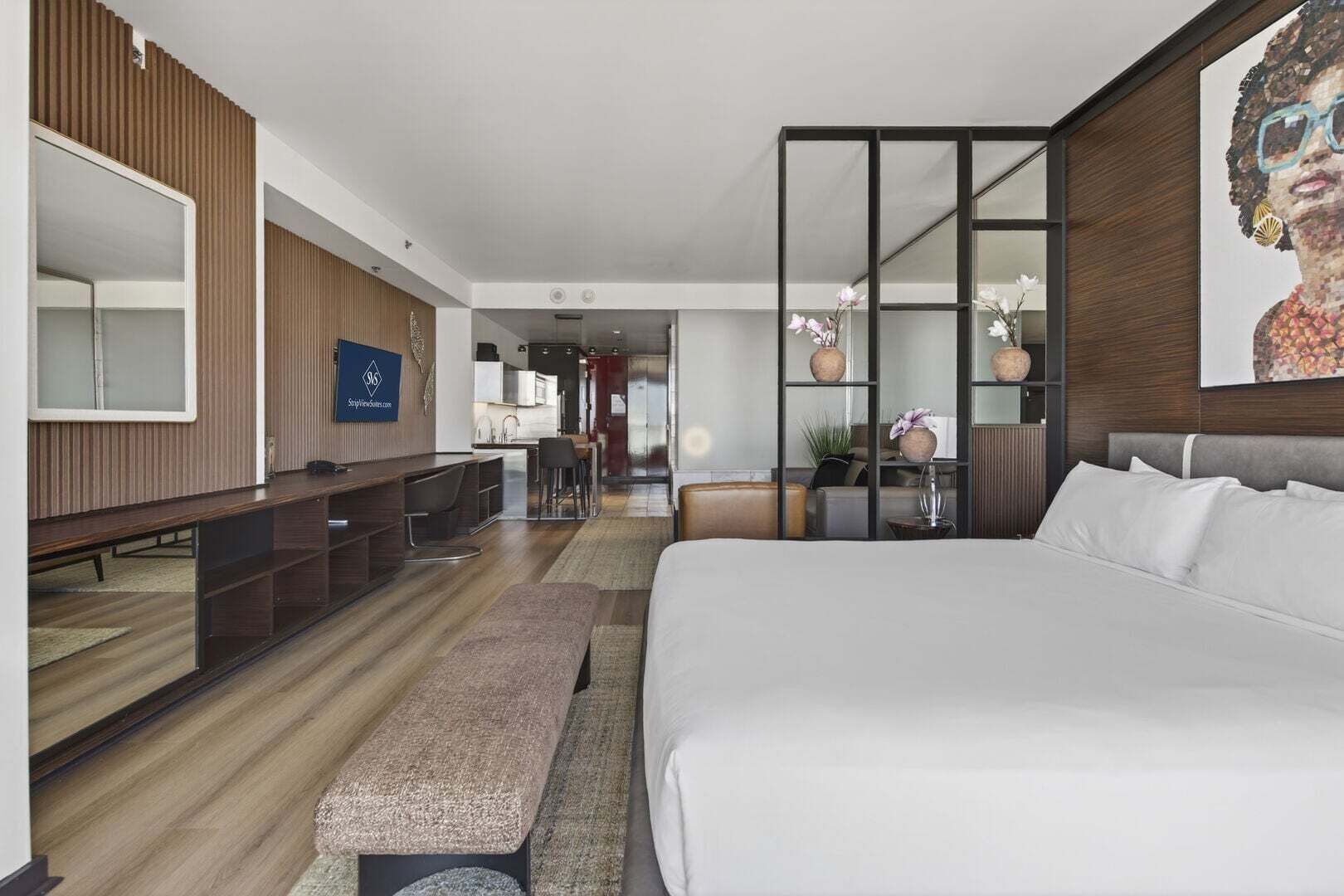 A modern hotel room with a large bed, a desk, a seating area, and a view into the kitchen area.
