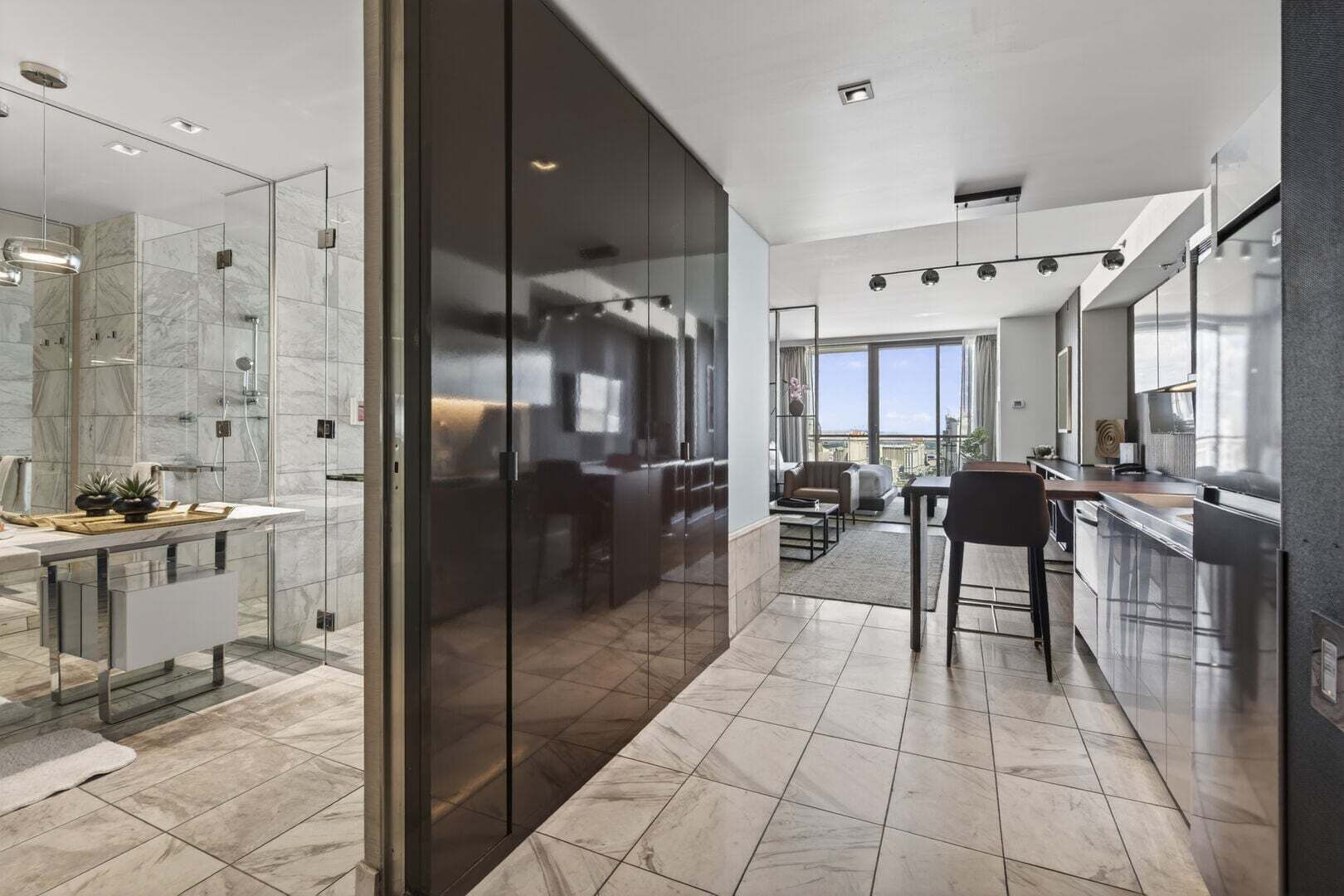 A modern hotel room with a glass partition, a bathroom with a shower, a dining area with a table and chairs, and a kitchen with stainless steel appliances.