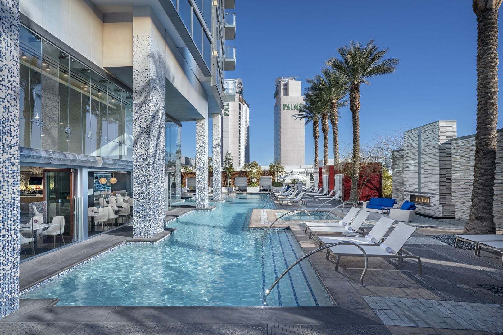 A hotel pool area with lounge chairs, a bar, and a tall building labeled 'PALMS' in the background.