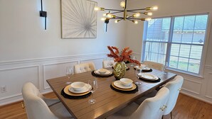 A dining room with a wooden table set for six, white upholstered chairs, a modern chandelier, and a large window with blinds. - GHome, Modern Cozy 5BR 10BEDS located  in Lilburn, GA with AC perfect for family (Lilburn)