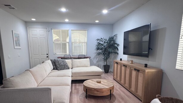 Ein Wohnzimmer mit einem Ecksofa, einem Couchtisch aus Holz, einem Fernseher auf einer Holzkommode und einer Topfpflanze.