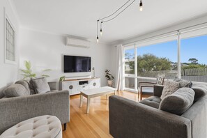 Ein modernes Wohnzimmer mit einem Flachbildfernseher, grauen Sofas, einem weißen Couchtisch und großen Fenstern mit Blick ins Freie.