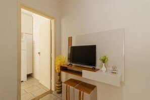 A modern room with a flat-screen TV mounted on the wall, a small wooden shelf below it, a vase with flowers, and a ship model. - Apartamento Pet Friendly Beira-Mar no Guilhermina (Praia Grande)